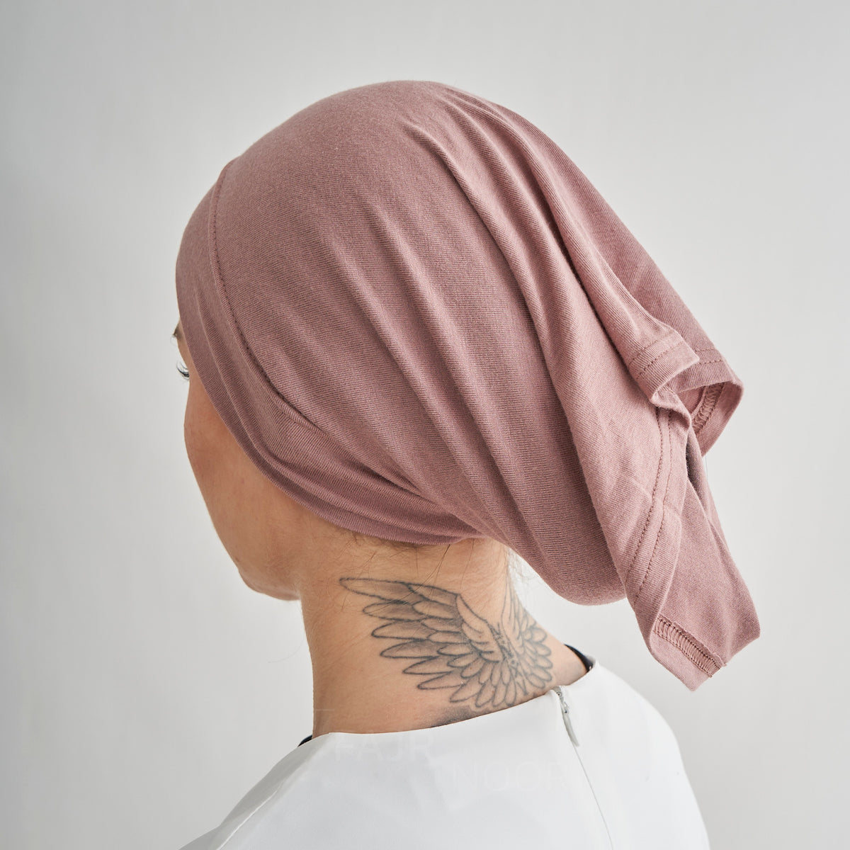 A person shown from the back wearing a dusty rose pink fabric head covering or hijab, with a wing tattoo visible on the neck, against a plain light gray background.