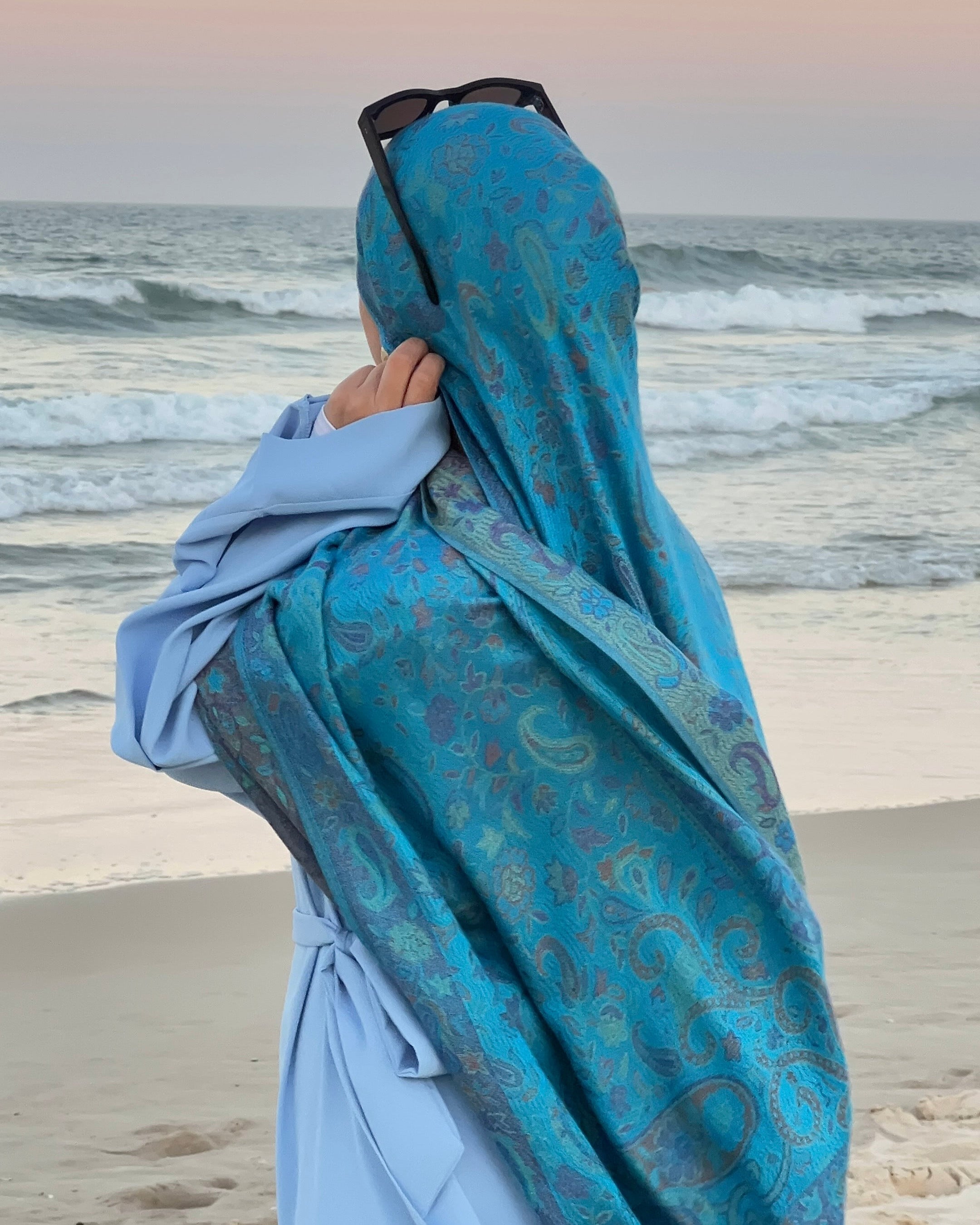 Person wearing a blue patterned hijab and sunglasses on a beach with ocean waves in the background