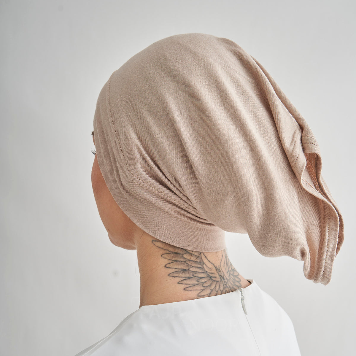 A person shown from the back wearing a soft, draped knit head covering in a light taupe-beige color. The shot is cropped from the side/back view, showing the fabric drape and texture clearly against a plain light gray background.