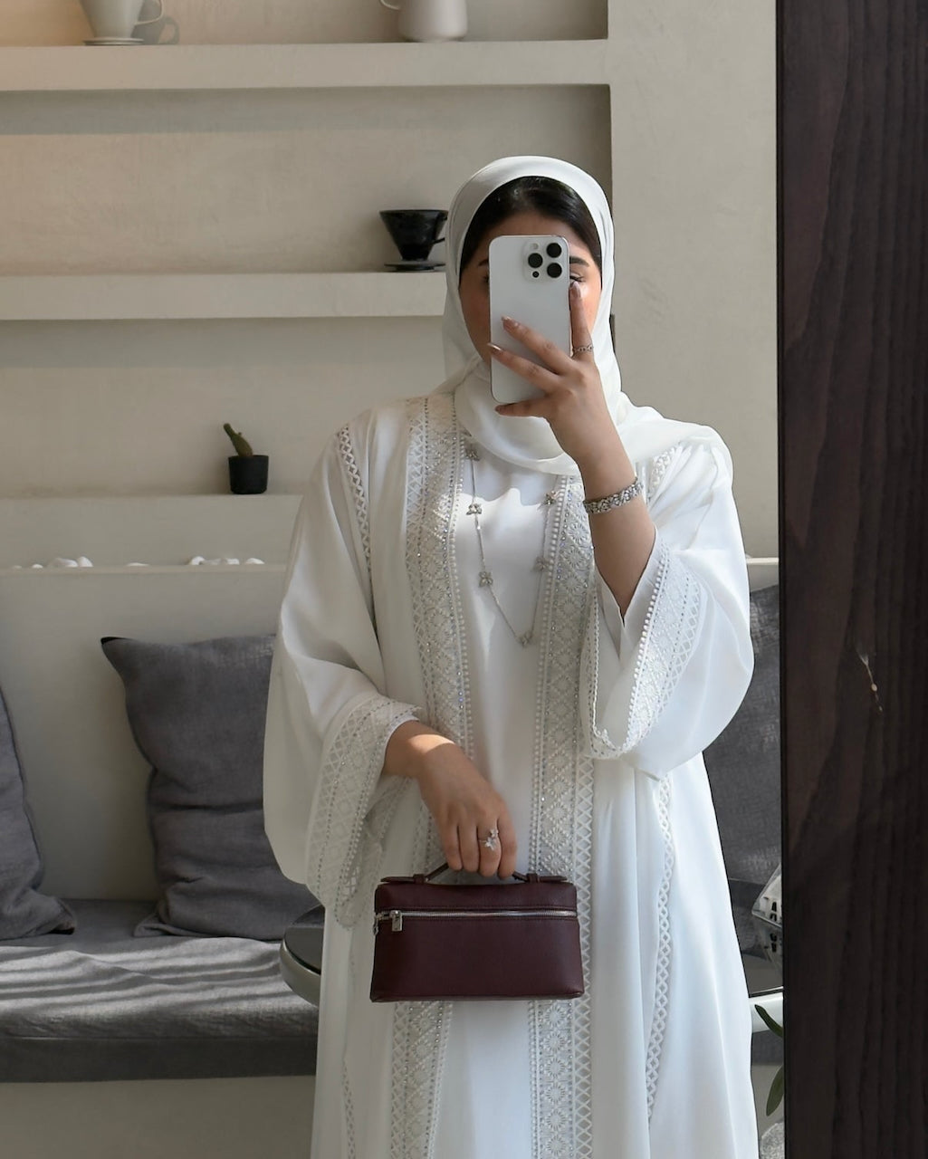 A woman takes a mirror selfie wearing a white embroidered abaya and matching headscarf, holding a dark burgundy handbag in a modern indoor setting. Nourah White Lace Abaya Modest Fashion - Fajr Noor