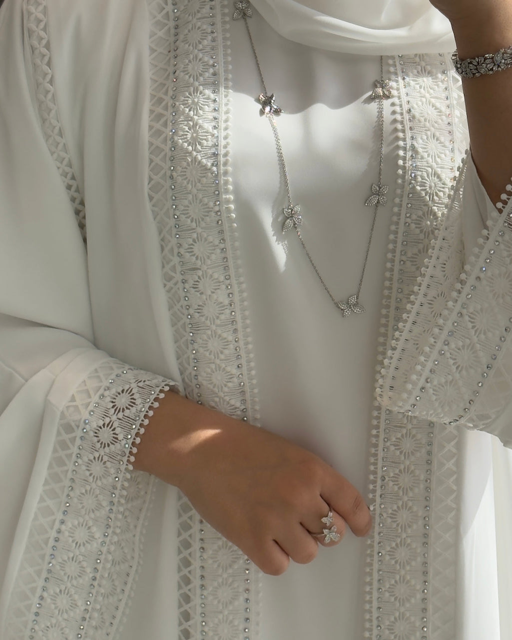 A close-up of a person wearing a white embroidered abaya or dress with silver-toned jewelry, including a necklace, bracelet, and ring, in soft natural light. Modest Fashion - Fajr Noor
