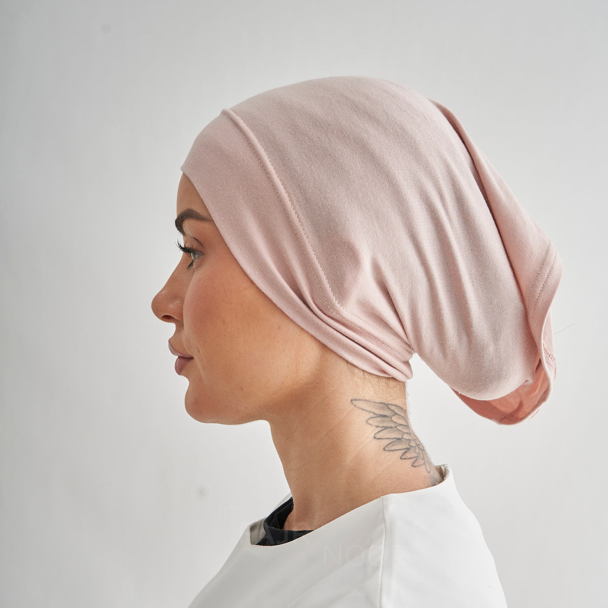 A woman in profile wearing a soft blush pink fabric head covering or hijab, shown against a plain light gray background.