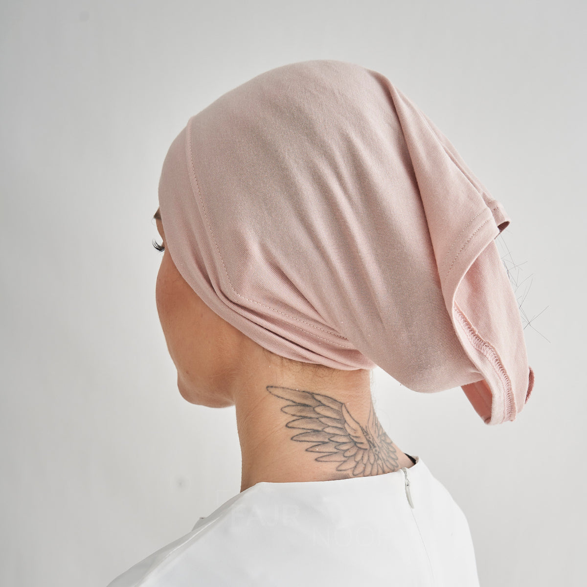 A person shown from the back wearing a soft blush pink fabric head covering or hijab, photographed against a plain light gray background.