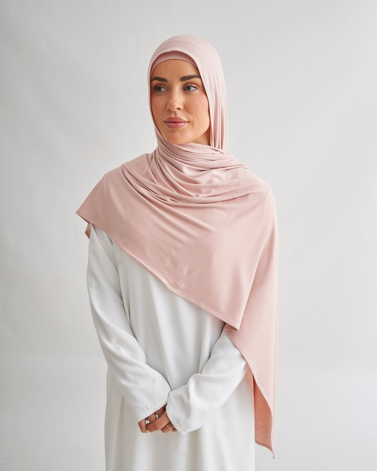 A woman wearing a nude pink hijab and a white long-sleeve dress, standing against a light gray studio background.