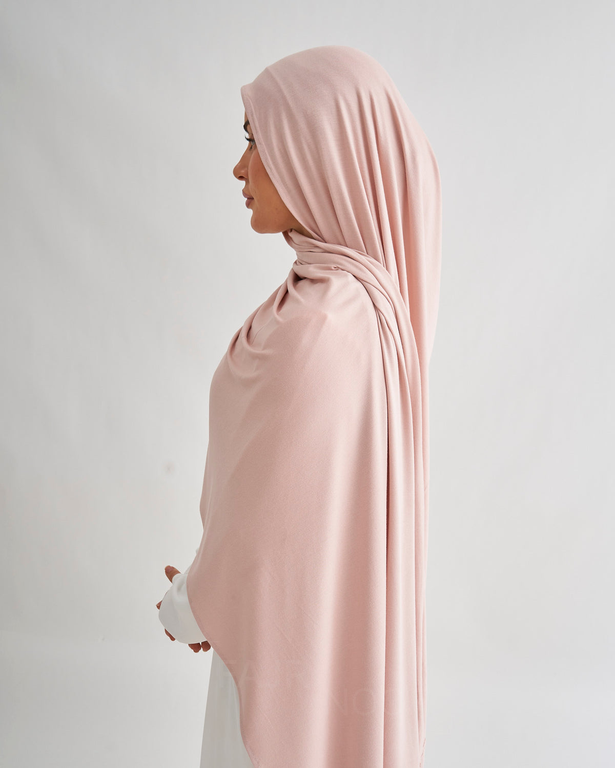 A woman in profile wearing a draped nude pink hijab against a light gray studio background.