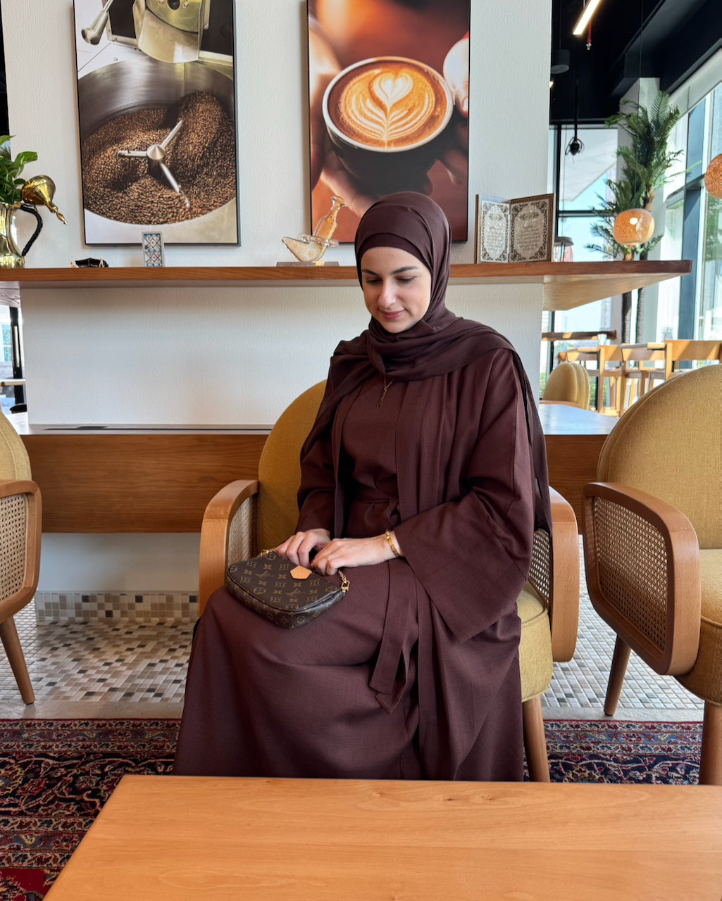 A woman wearing a dark brown coffee linen abaya and matching hijab sits in a cafe chair, holding a patterned clutch purse. The background shows a cozy cafe interior with wooden furniture, a patterned rug, and coffee-themed wall art.