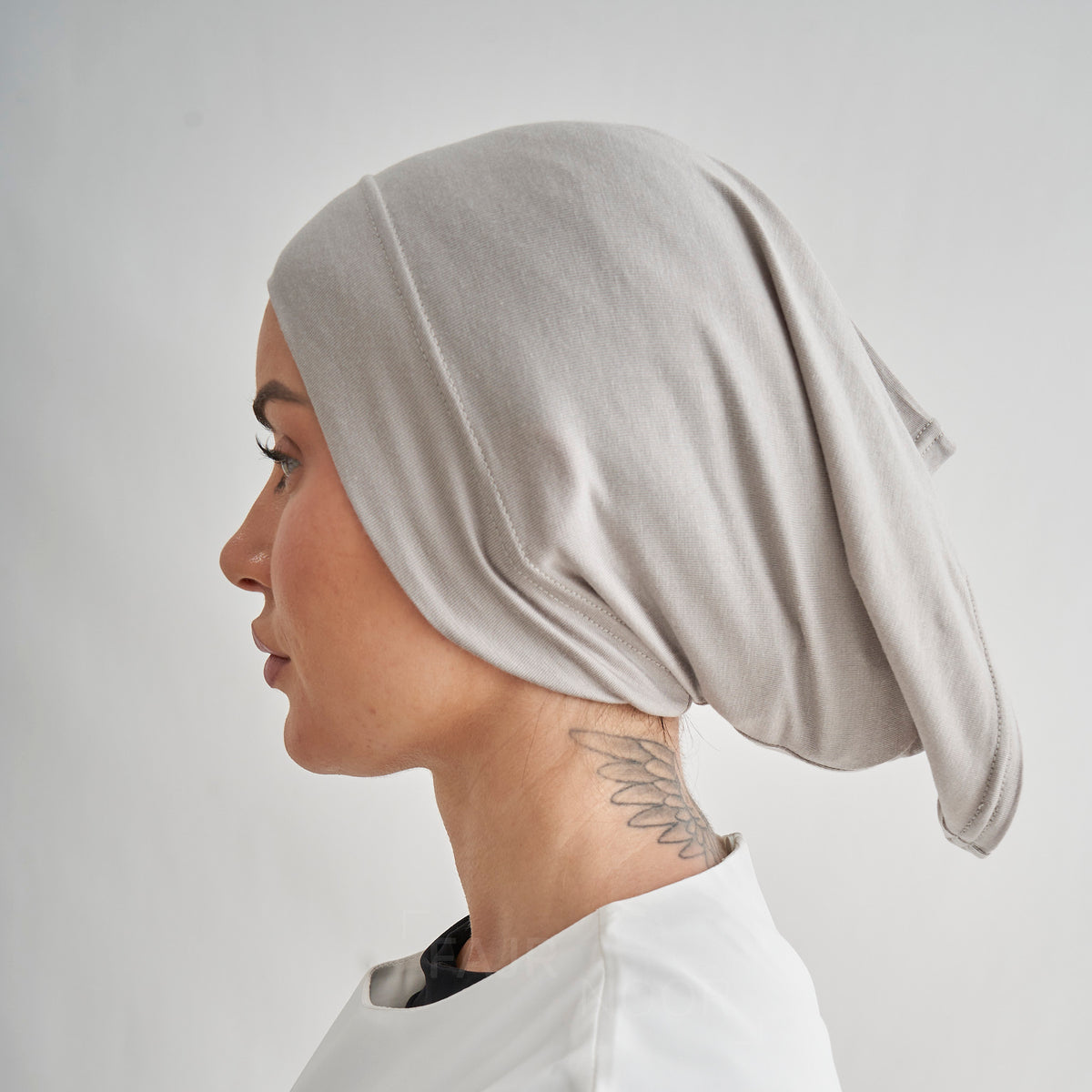 A woman shown in profile wearing a soft, draped light beige or taupe fabric head covering. The shot is taken against a plain light gray studio background.