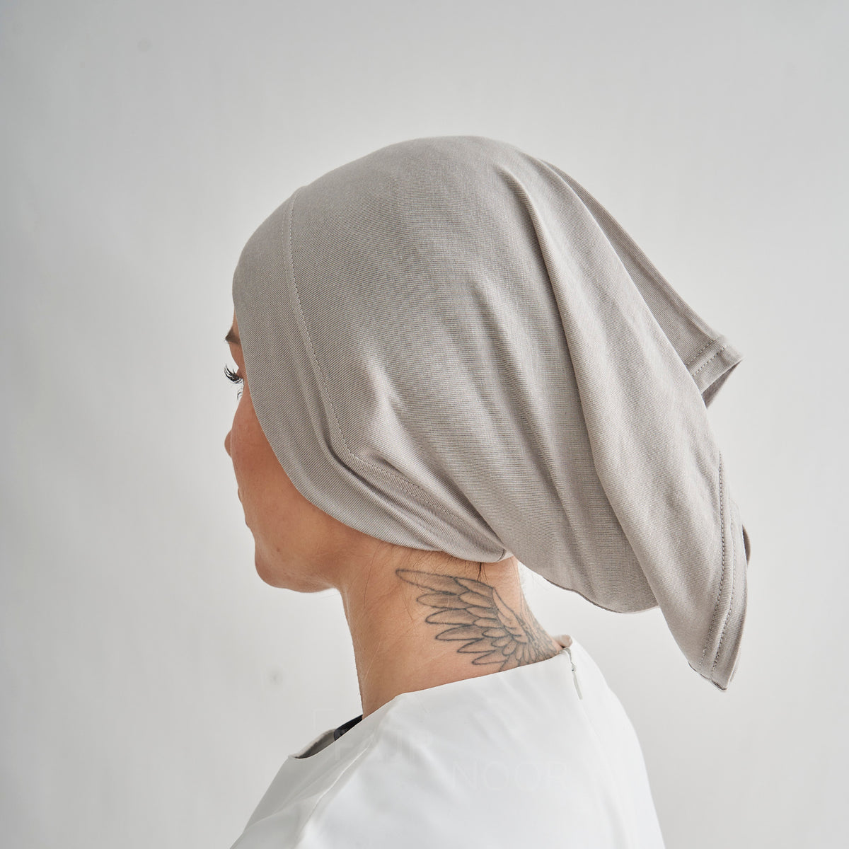 A person shown in side profile wearing a light taupe-beige textured fabric head covering or wrap, photographed against a plain light gray background.