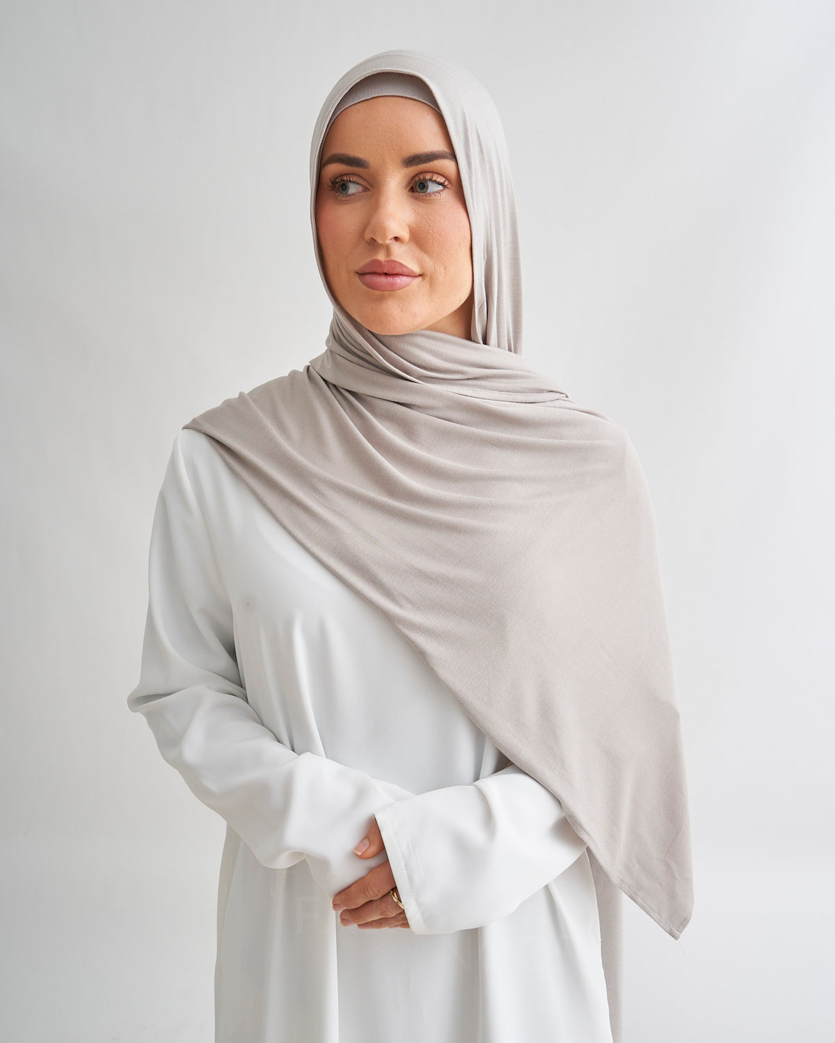 A woman wearing a light taupe-beige hijab and a white long-sleeve abaya, standing against a light gray studio background.