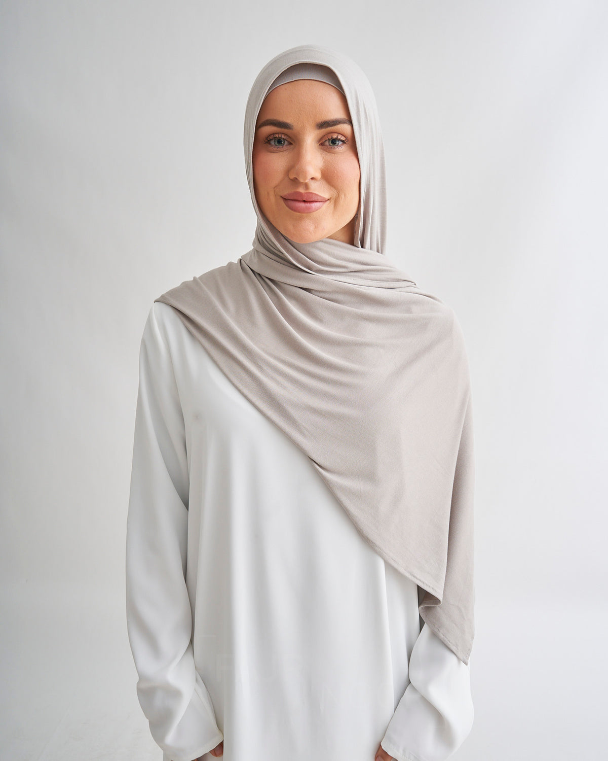 A woman facing forward wearing a light taupe draped hijab and a white long-sleeve top against a light gray studio background.