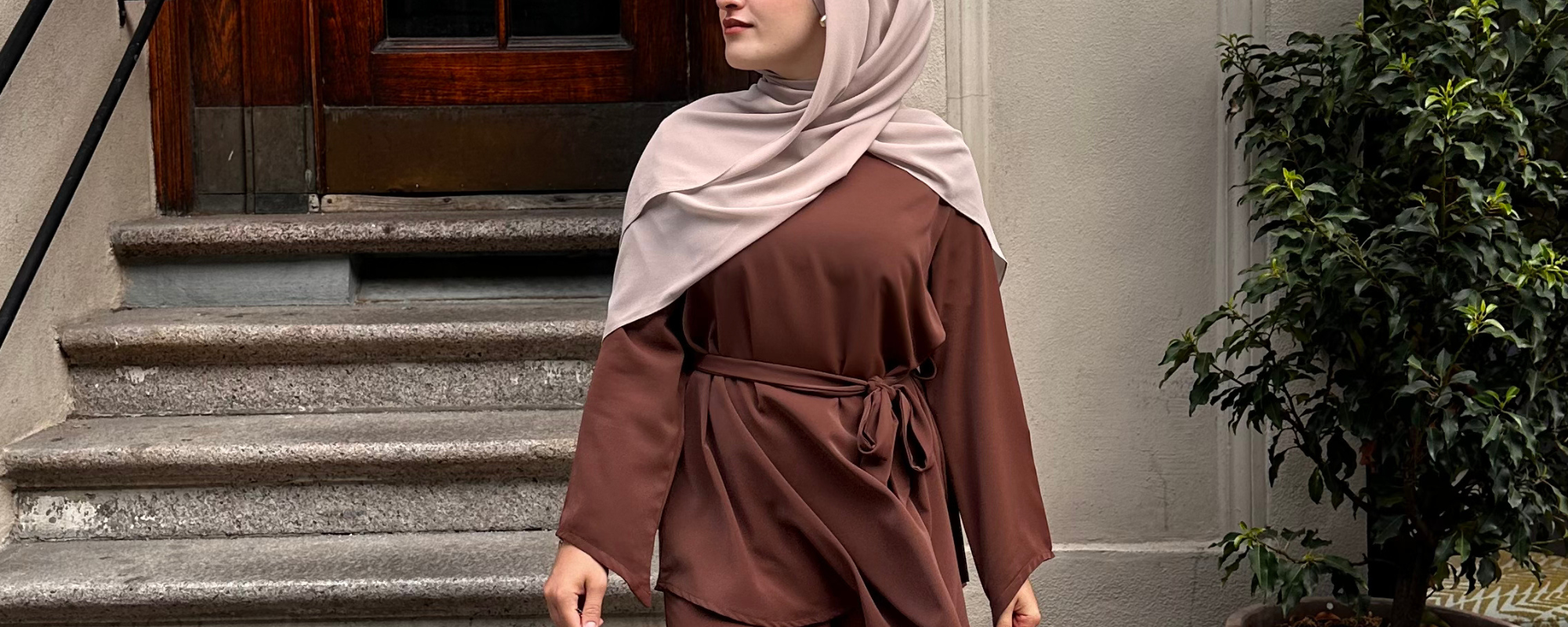 Woman in a brown outfit and beige hijab standing on stone steps with a plant in the background