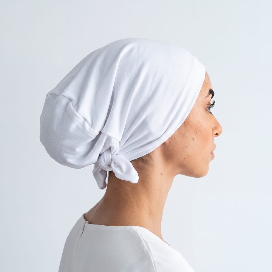 A woman shown in profile wearing a white fabric head covering tied at the back of her neck, against a plain light gray background.