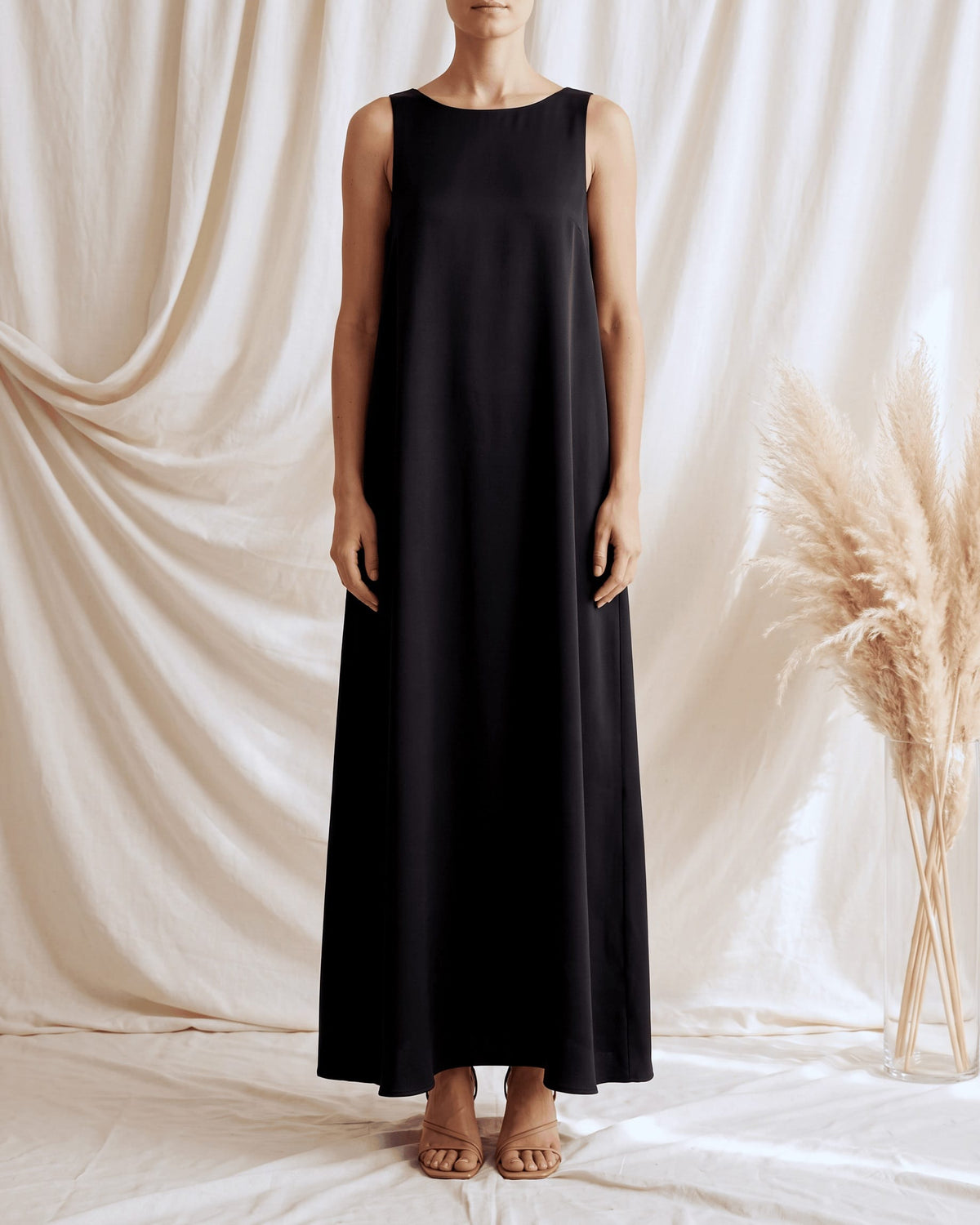 A sleeveless black maxi dress worn by a model standing against a draped cream fabric backdrop, with a vase of dried pampas grass on the right.