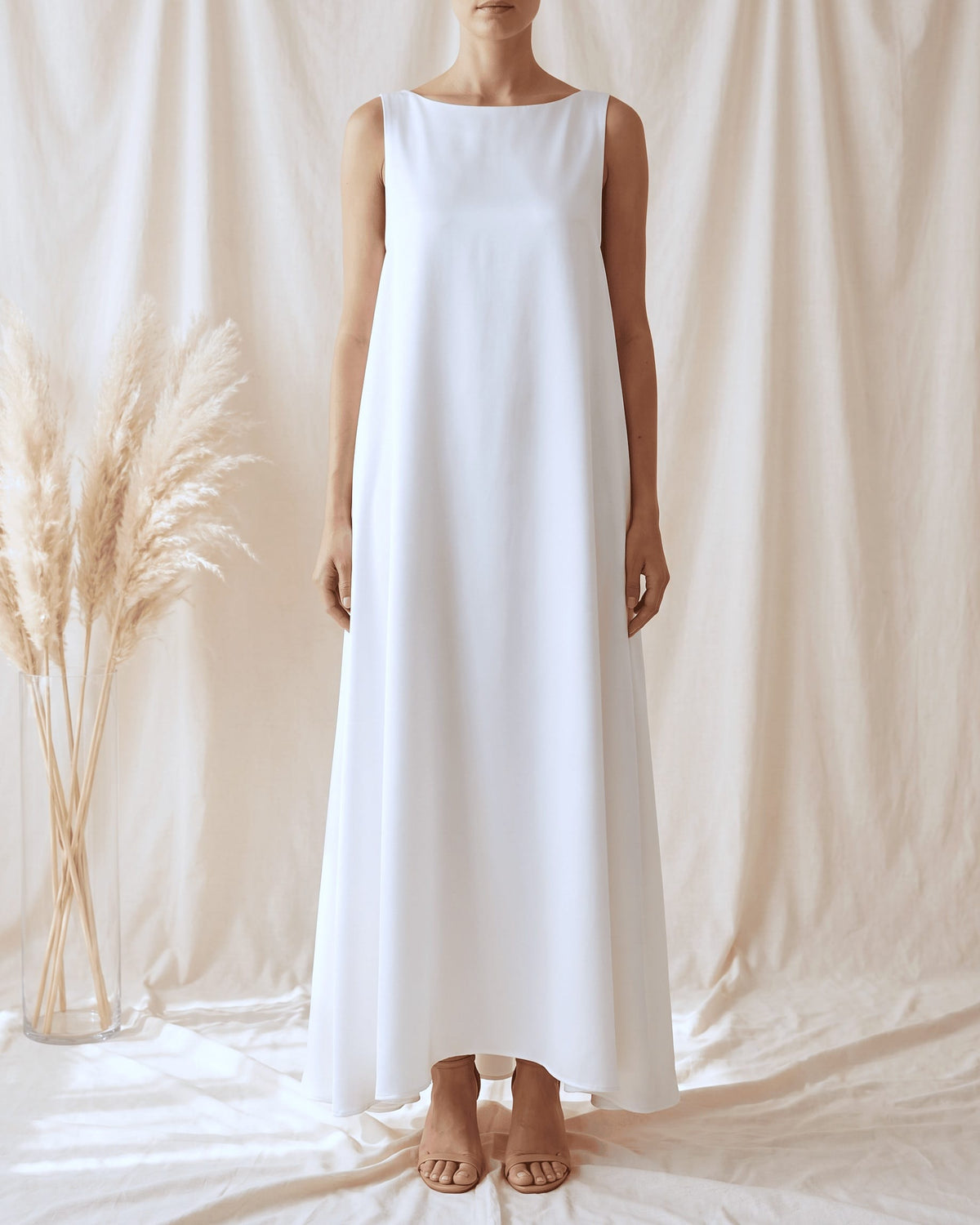 A woman wearing a sleeveless white maxi dress with a boat neckline, standing against a draped cream fabric backdrop with dried pampas grass on the left.