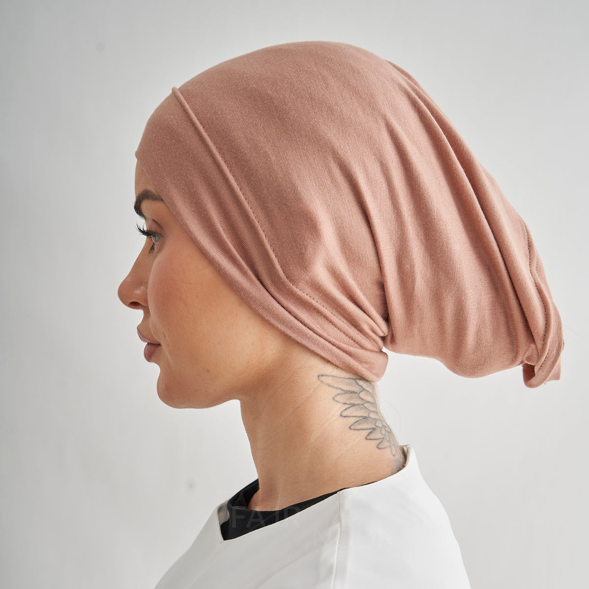 A woman shown in profile wearing a soft, draped dusty rose head covering or hijab against a plain light gray background.