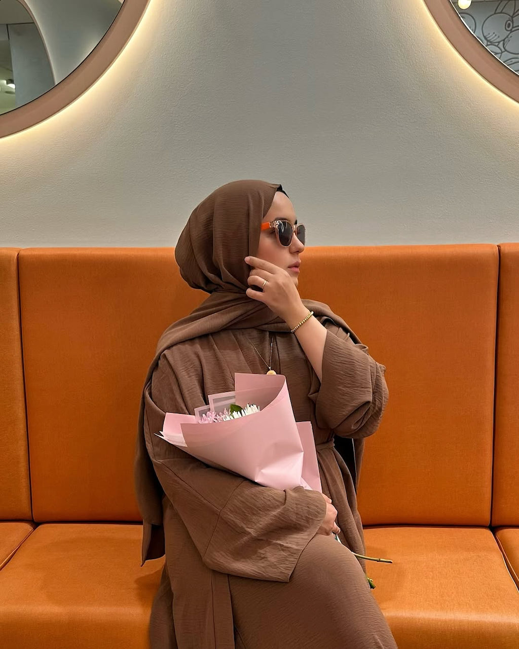 A woman wearing a brown hijab and matching brown outfit sits on an orange bench holding a pink wrapped bouquet. She is wearing sunglasses and posing in a modern indoor setting with a curved wall and circular light fixture.