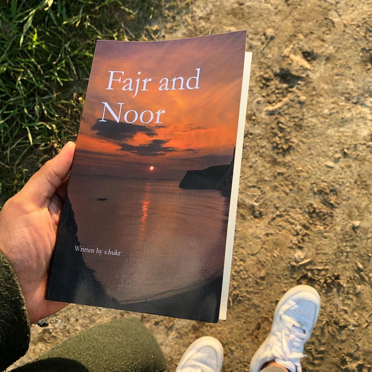 A hand holds a book titled 'Fajr and Noor' against an outdoor background of a dirt path and grass. The book cover features a sunset over a sea with cliffs, and the text 'Written by s.