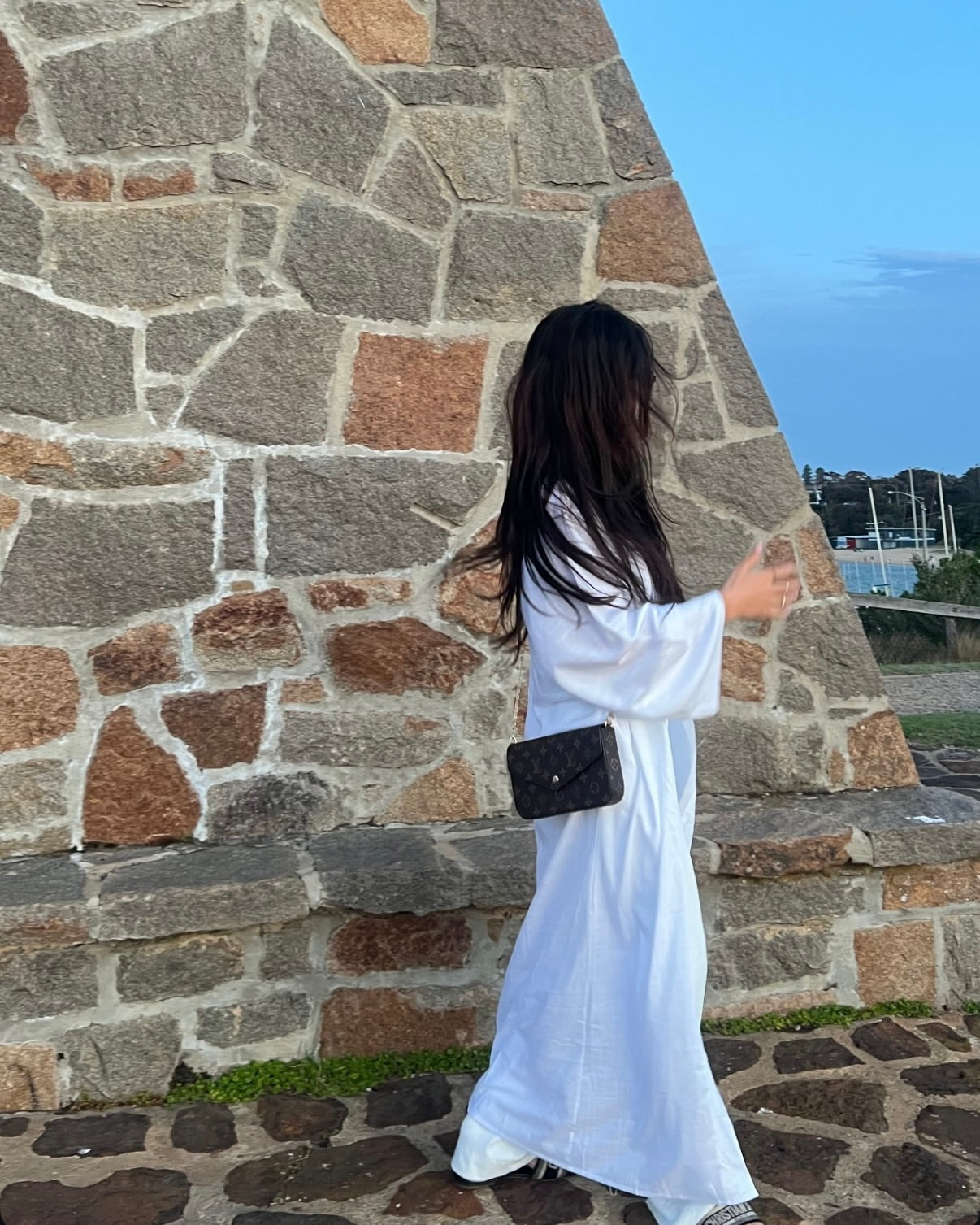 Person in a white dress standing against a stone wall with a clear sky.