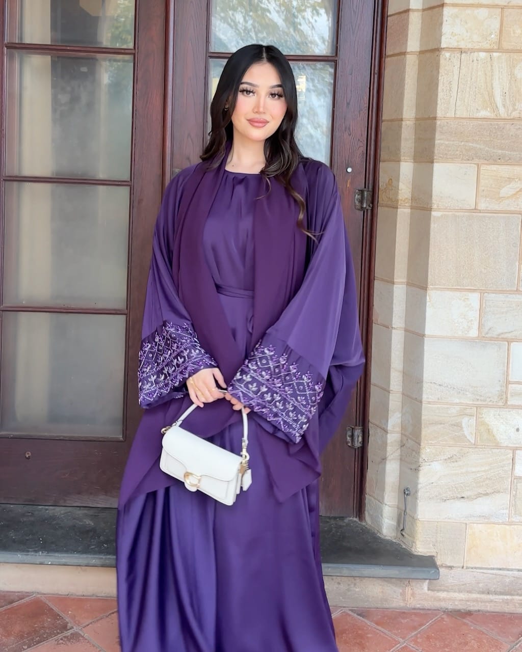 A woman wearing a flowing purple abaya dress with embroidered cuffs, holding a small white handbag, standing in front of a stone and wood door.