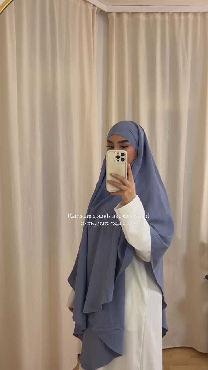 Woman wearing the Fajr Noor khimar in dusty blue over a white abaya, taking a mirror selfie