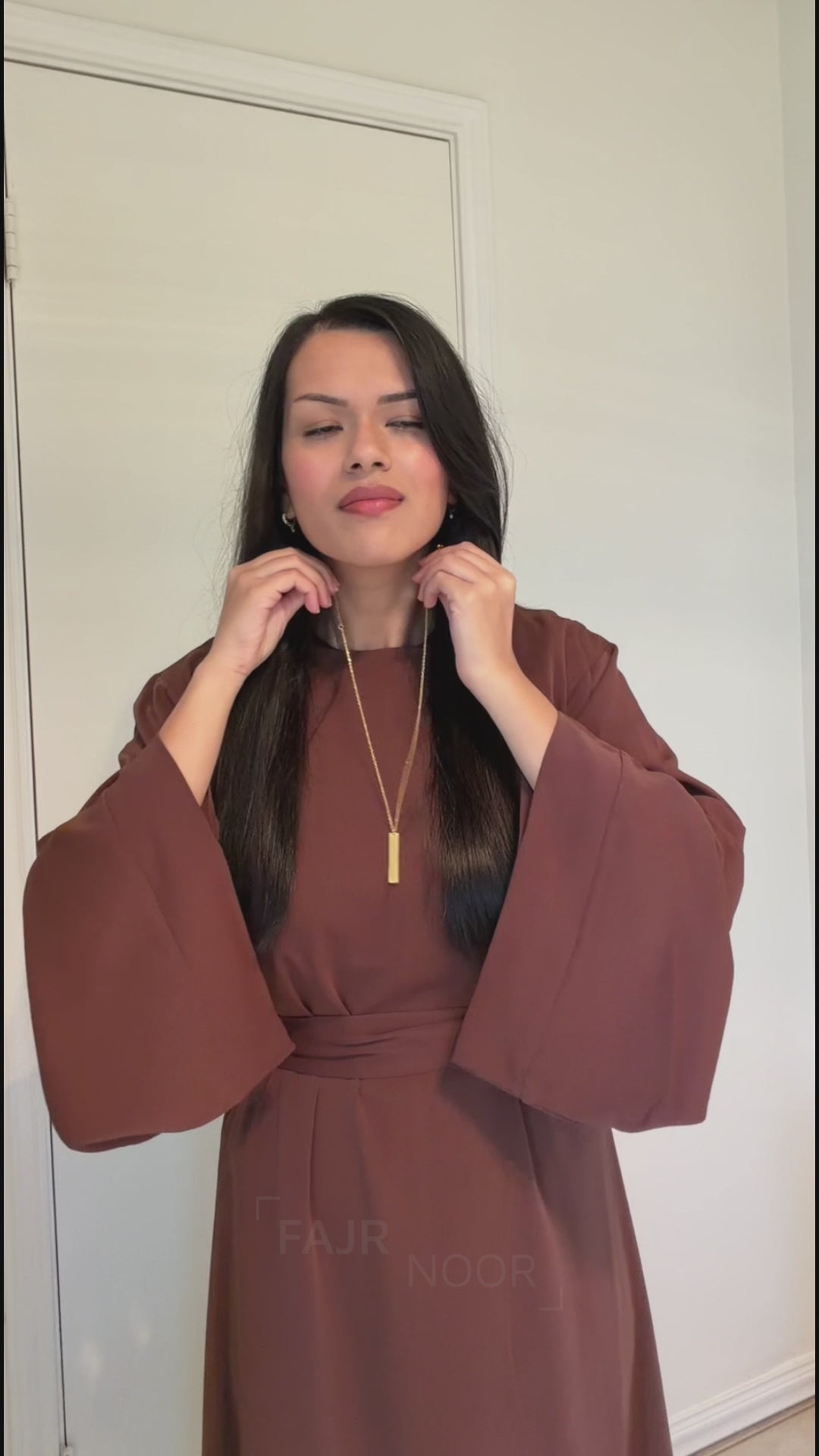 Woman wearing the Fajr Noor Khatijah Abaya in coffee with a gold "Verily, with hardship comes ease" bar necklace and Clover Bracelet