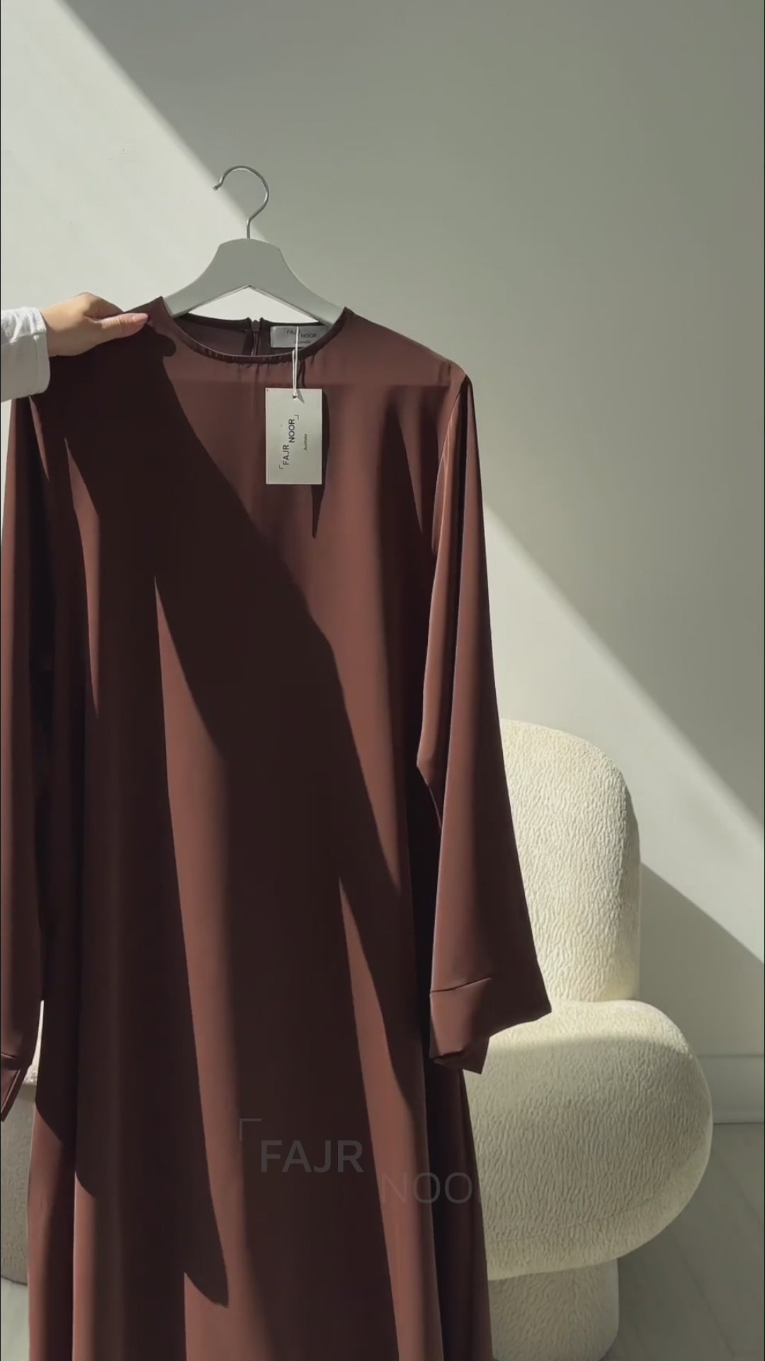 Woman wearing the Fajr Noor Khatijah Abaya in brown with a cream hijab, sitting and reading a book indoors