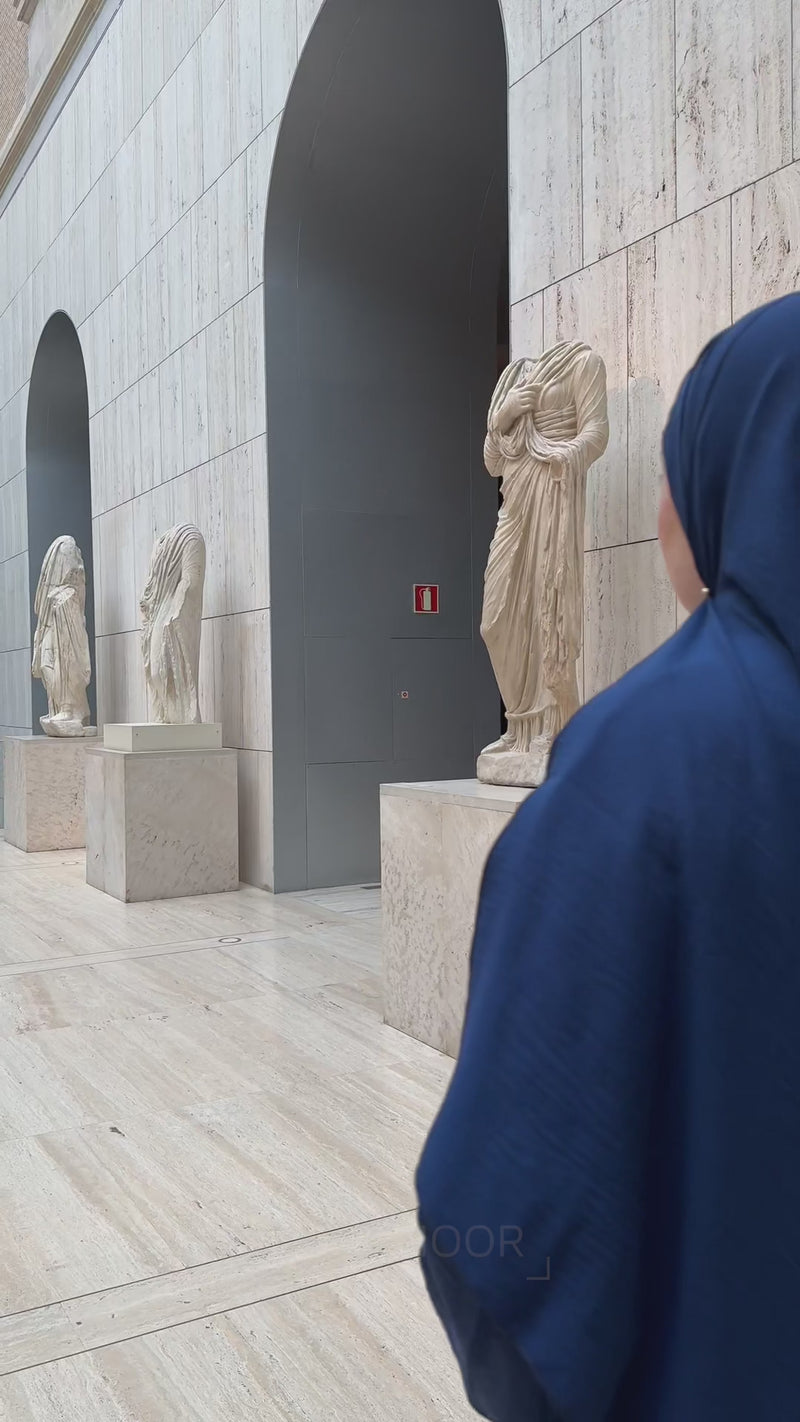 Woman wearing the Fajr Noor 3-piece Crinkle Abaya in navy with an Ayatul Kursi necklace, standing in a gallery