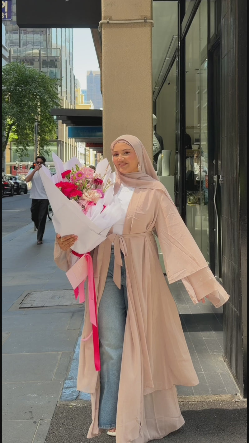 Woman wearing the Fajr Noor Aisha Abaya in nude with a matching hijab, holding a bouquet of flowers on a city street