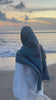 Woman wearing the Fajr Noor Khatijah Abaya in white with a forest green pashmina hijab and Ayatul Kursi necklace, on the beach at sunset