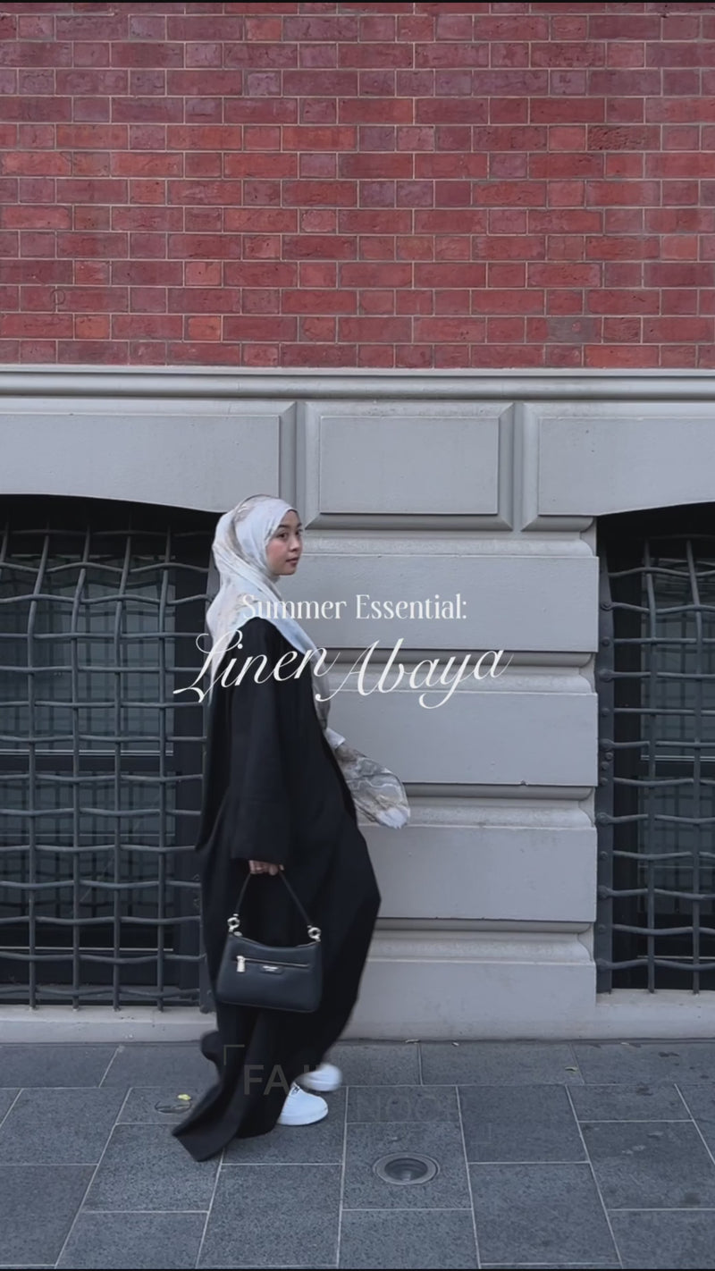 Woman wearing the Fajr Noor Aya Linen Abaya in black with a white hijab, walking outdoors as a summer essential