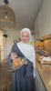 Woman wearing the Fajr Noor Khatijah Abaya in charcoal with a white hijab, enjoying cake in her kitchen