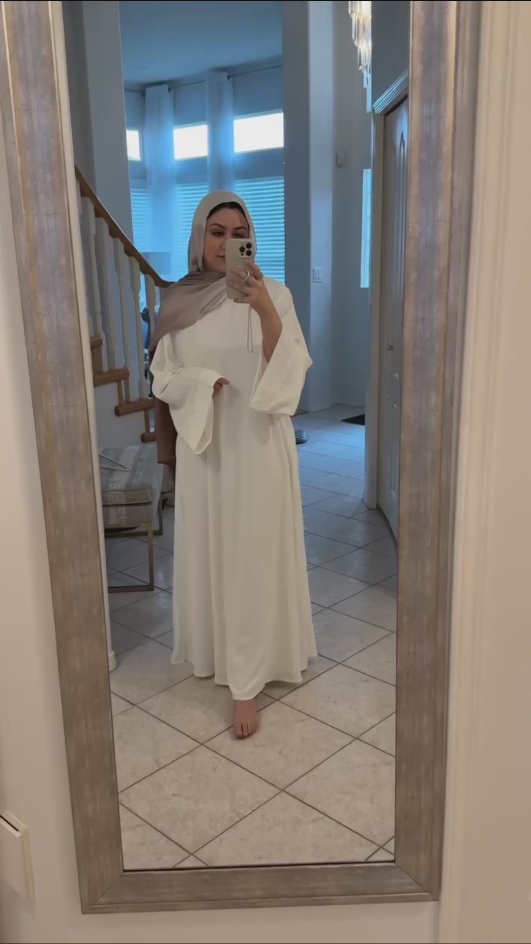 Woman wearing the Fajr Noor Khatijah Abaya in white with a taupe hijab, taking a mirror selfie at home