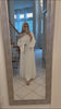 Woman wearing the Fajr Noor Khatijah Abaya in white with a taupe hijab, taking a mirror selfie at home