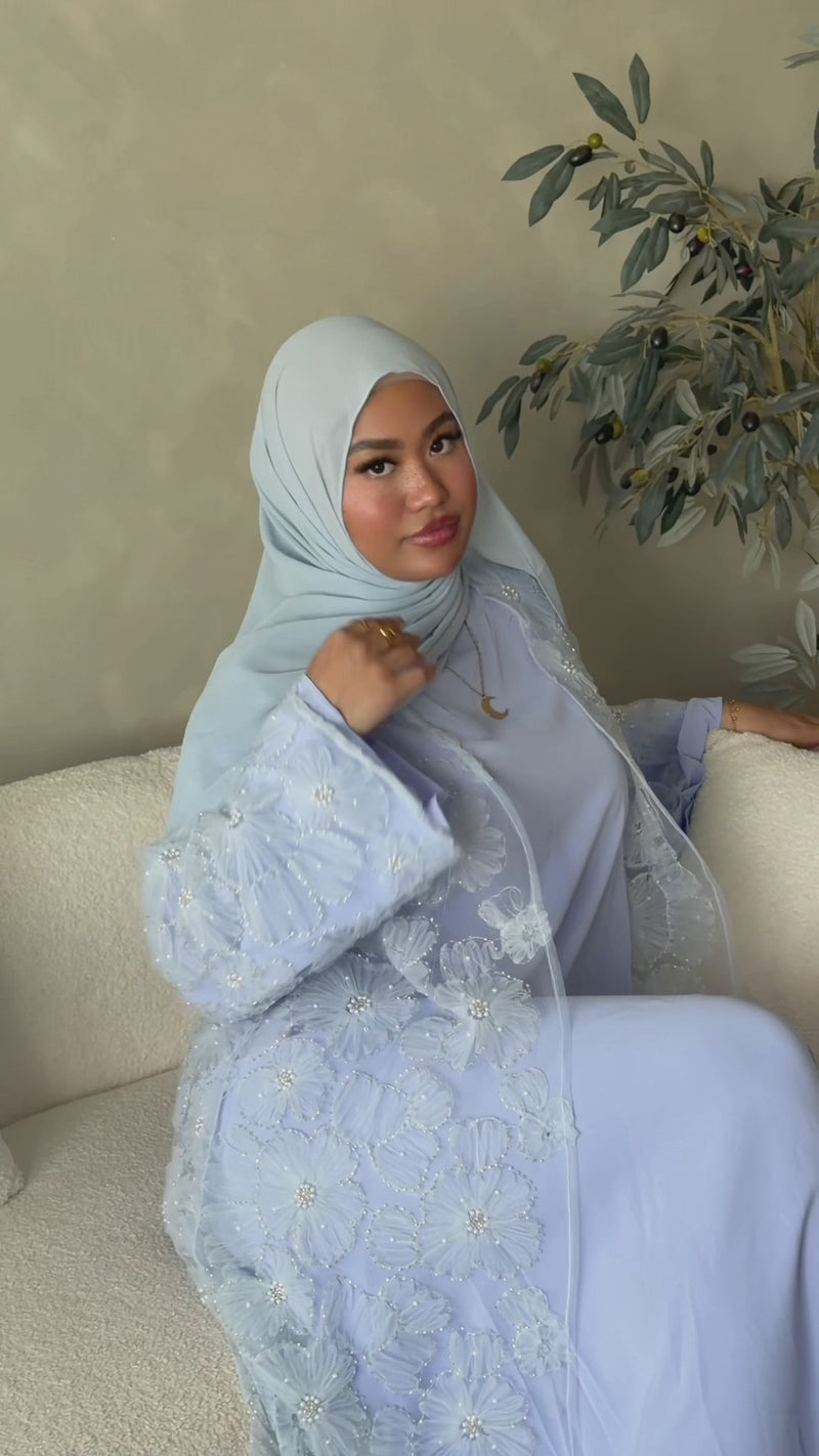 Woman seated wearing the Fajr Noor Sumayya Abaya in sky blue with 3D floral appliqué detail, matching hijab
