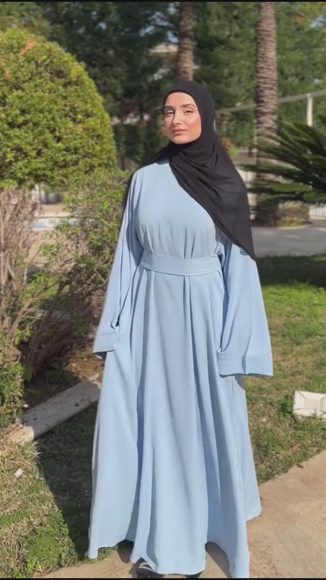 Woman wearing the Fajr Noor Khatijah Abaya in sky blue with a berry hijab, walking outdoors near palm trees