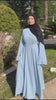 Woman wearing the Fajr Noor Khatijah Abaya in sky blue with a berry hijab, walking outdoors near palm trees