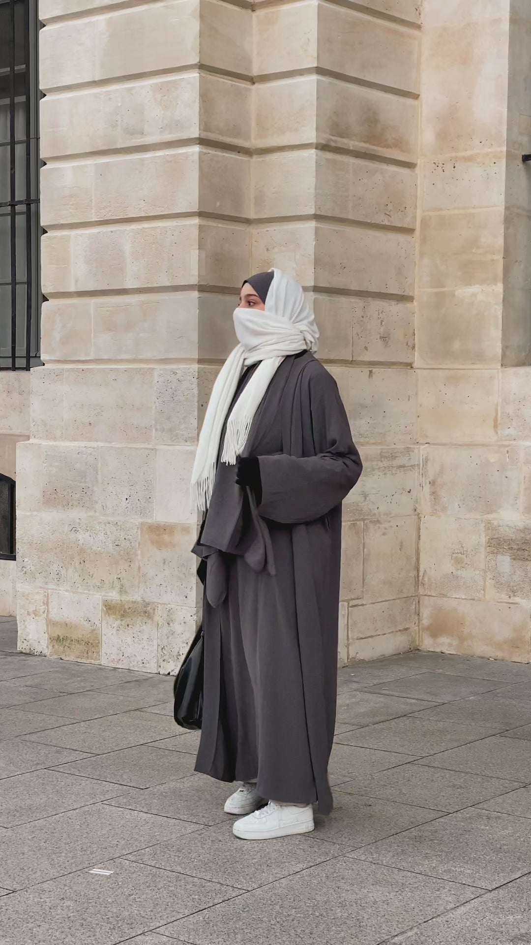 Woman wearing the Fajr Noor Crinkle Abaya in grey with a white hijab, walking past stone architecture, styled with sneakers