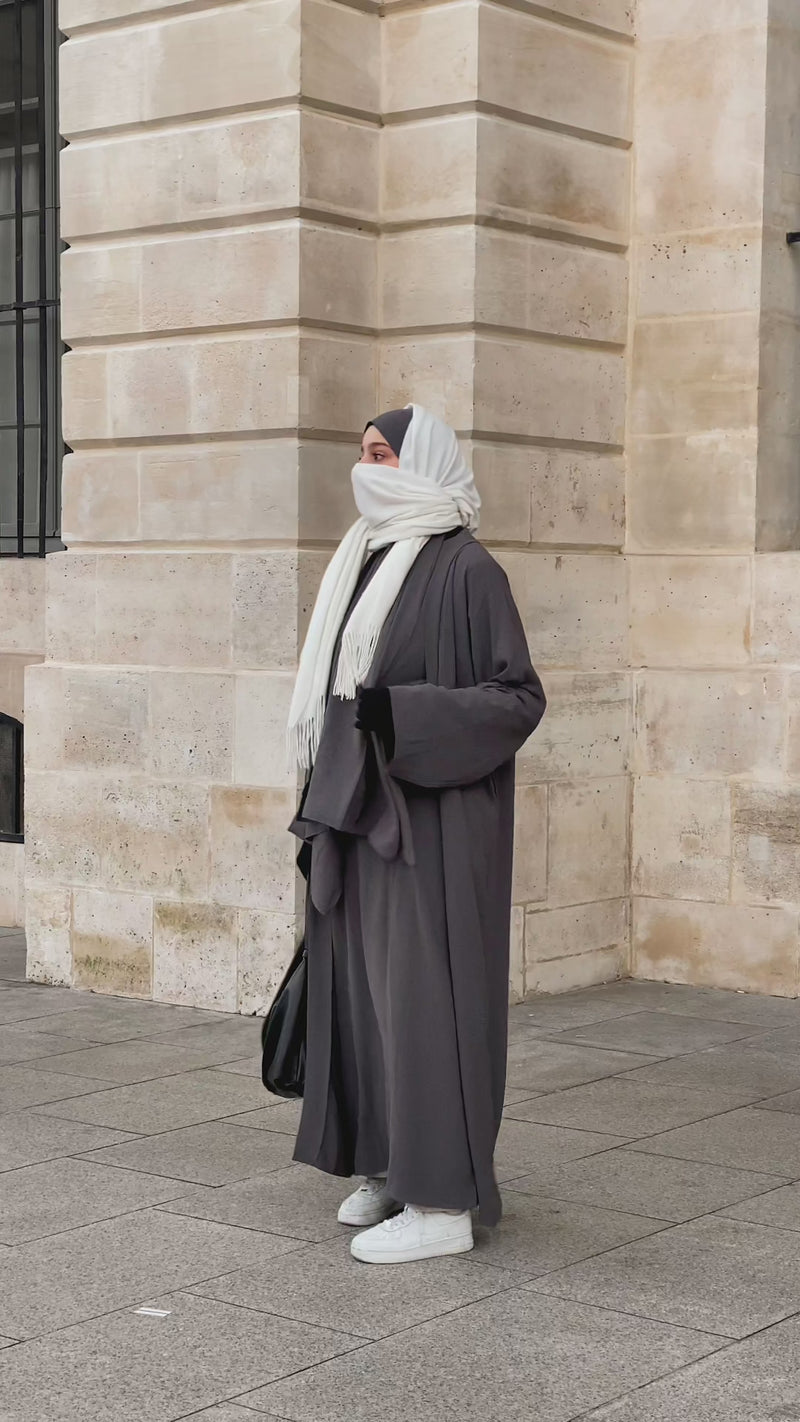 Woman wearing the Fajr Noor Crinkle Abaya in grey with a white hijab, walking past stone architecture, styled with sneakers