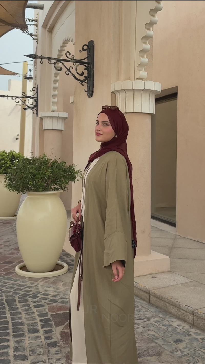Woman wearing the Fajr Noor Aya Linen Abaya in khaki with a maroon hijab, walking outdoors near an archway
