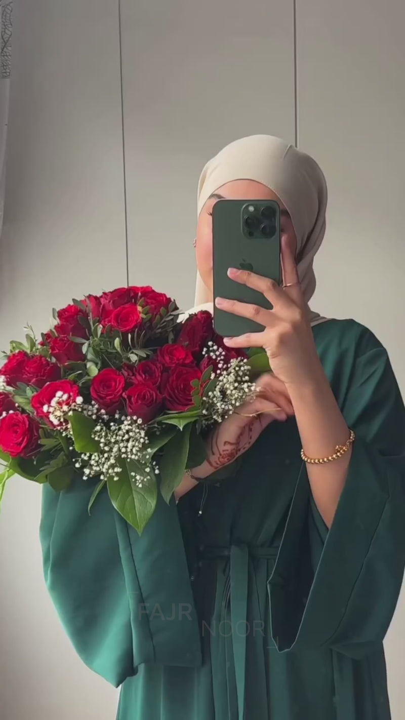 Woman wearing the Fajr Noor Khatijah Abaya in emerald with a taupe hijab, holding a bouquet of red roses