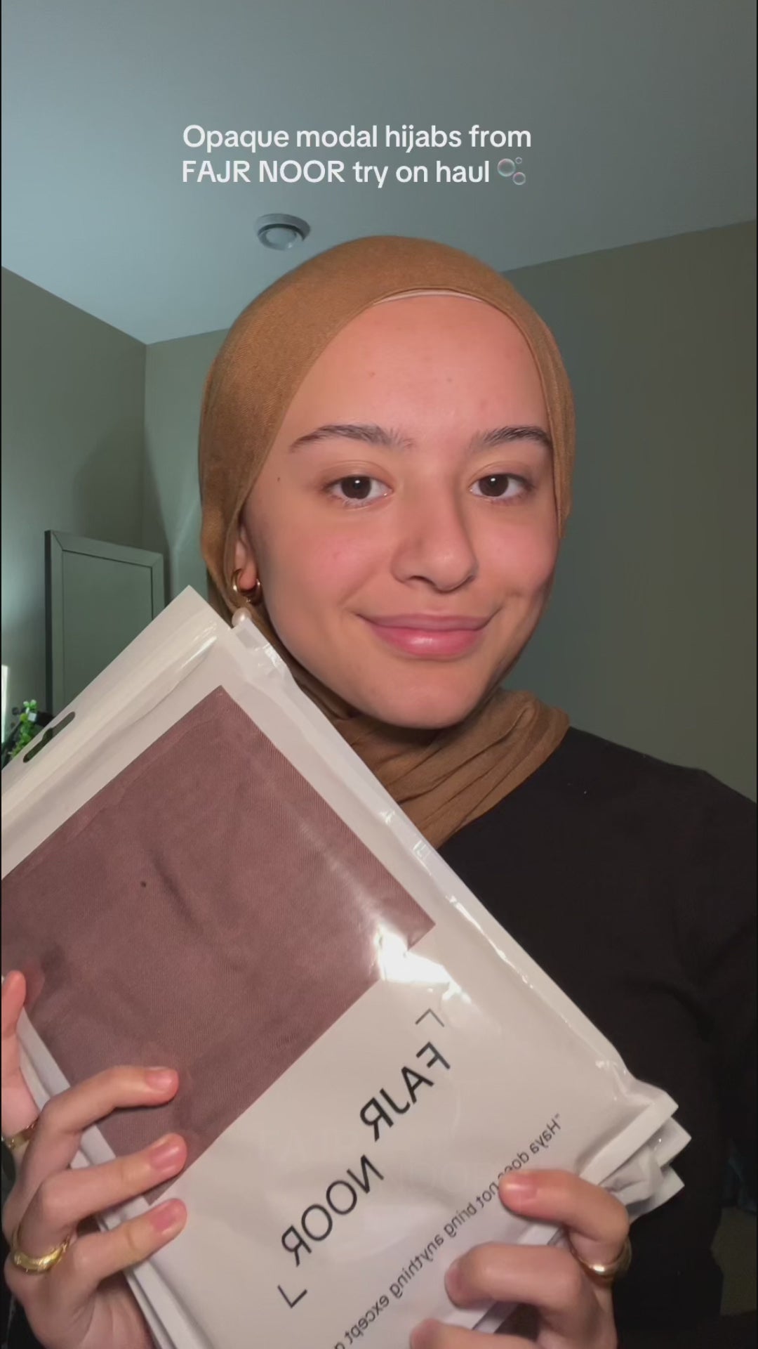 Woman wearing a mustard gold Fajr Noor opaque premium modal hijab, holding packaged modal hijabs during a try-on haul