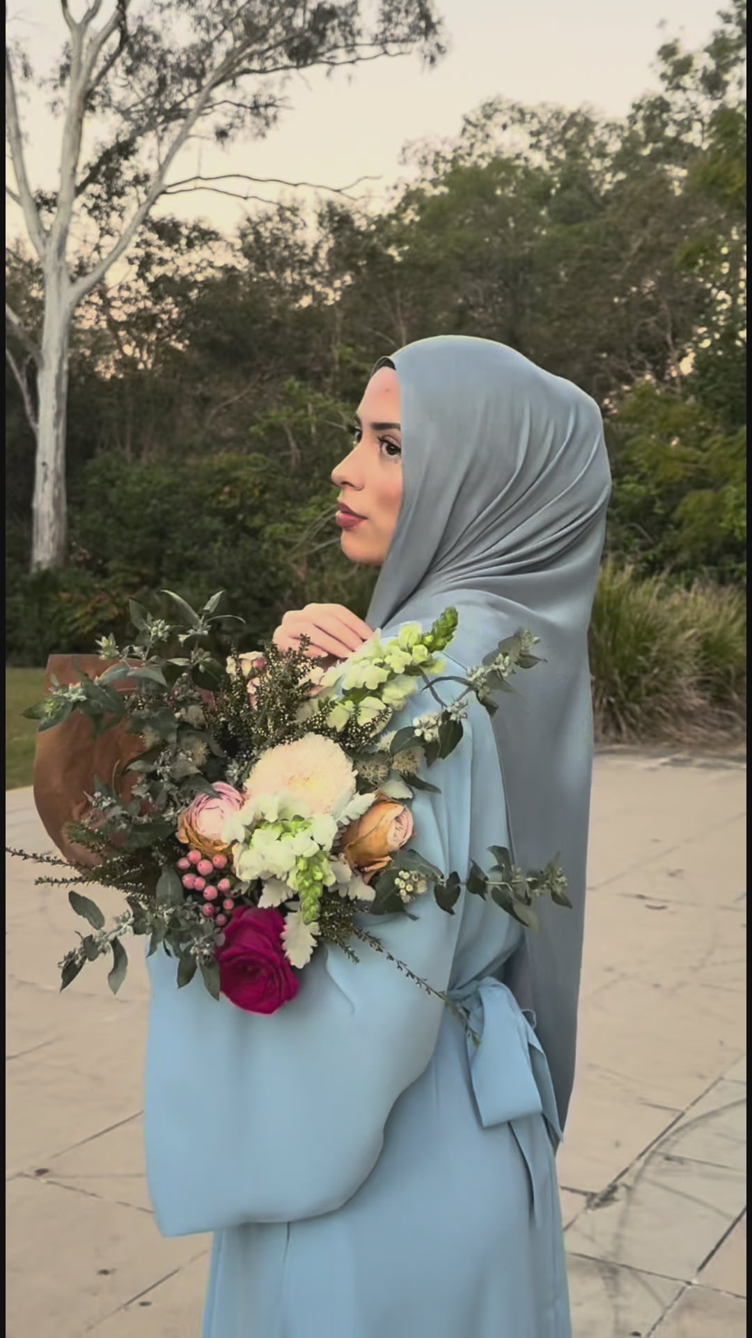 Woman wearing the Fajr Noor Khatijah Abaya in sky blue with a matching modal hijab, holding a floral bouquet outdoors