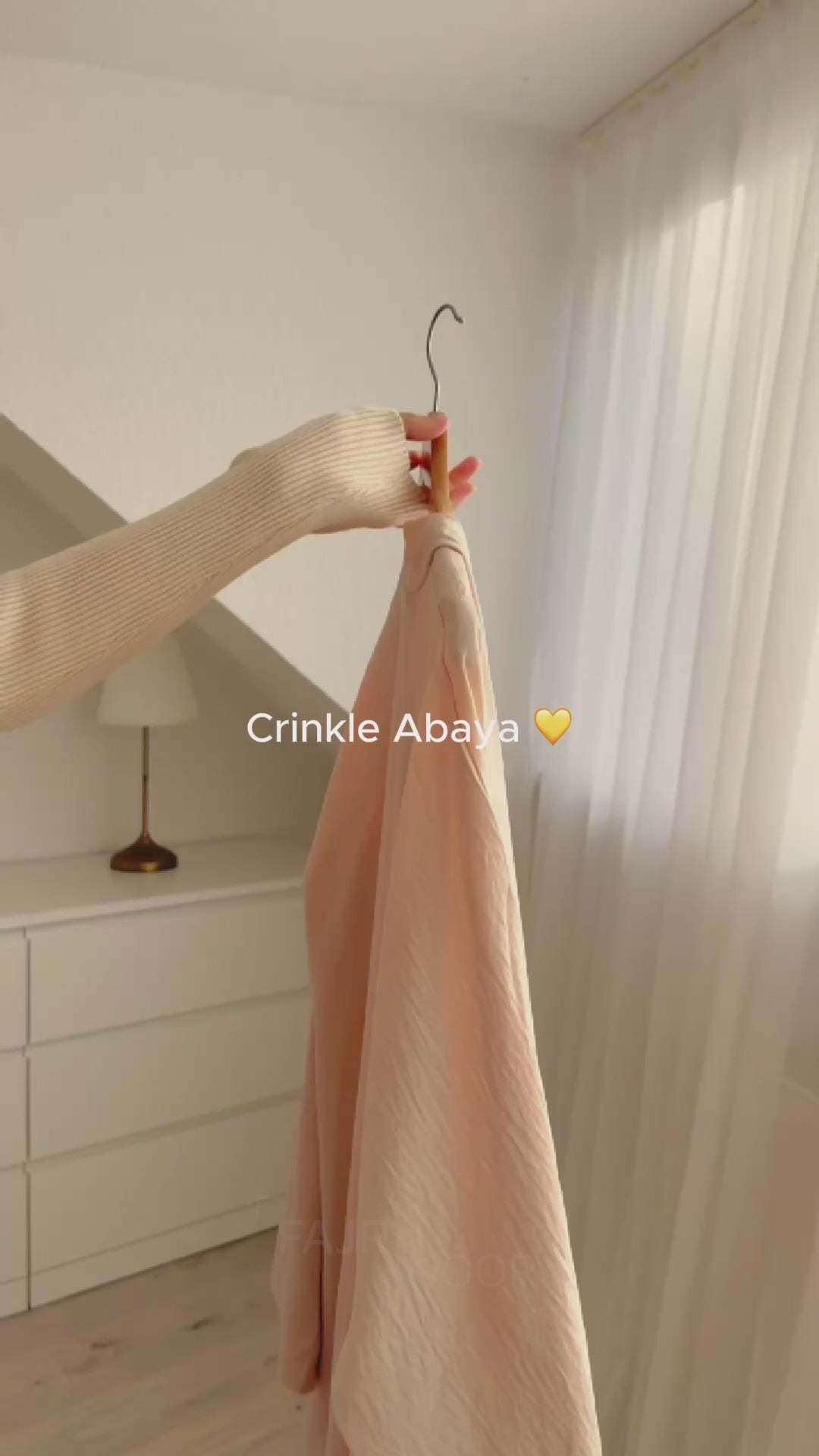 Fajr Noor Crinkle Abaya in cream held on a hanger by a window, showing the flowing crinkle texture