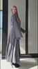 Woman wearing the Fajr Noor Khatijah Abaya in charcoal with a tan hijab, standing by a window