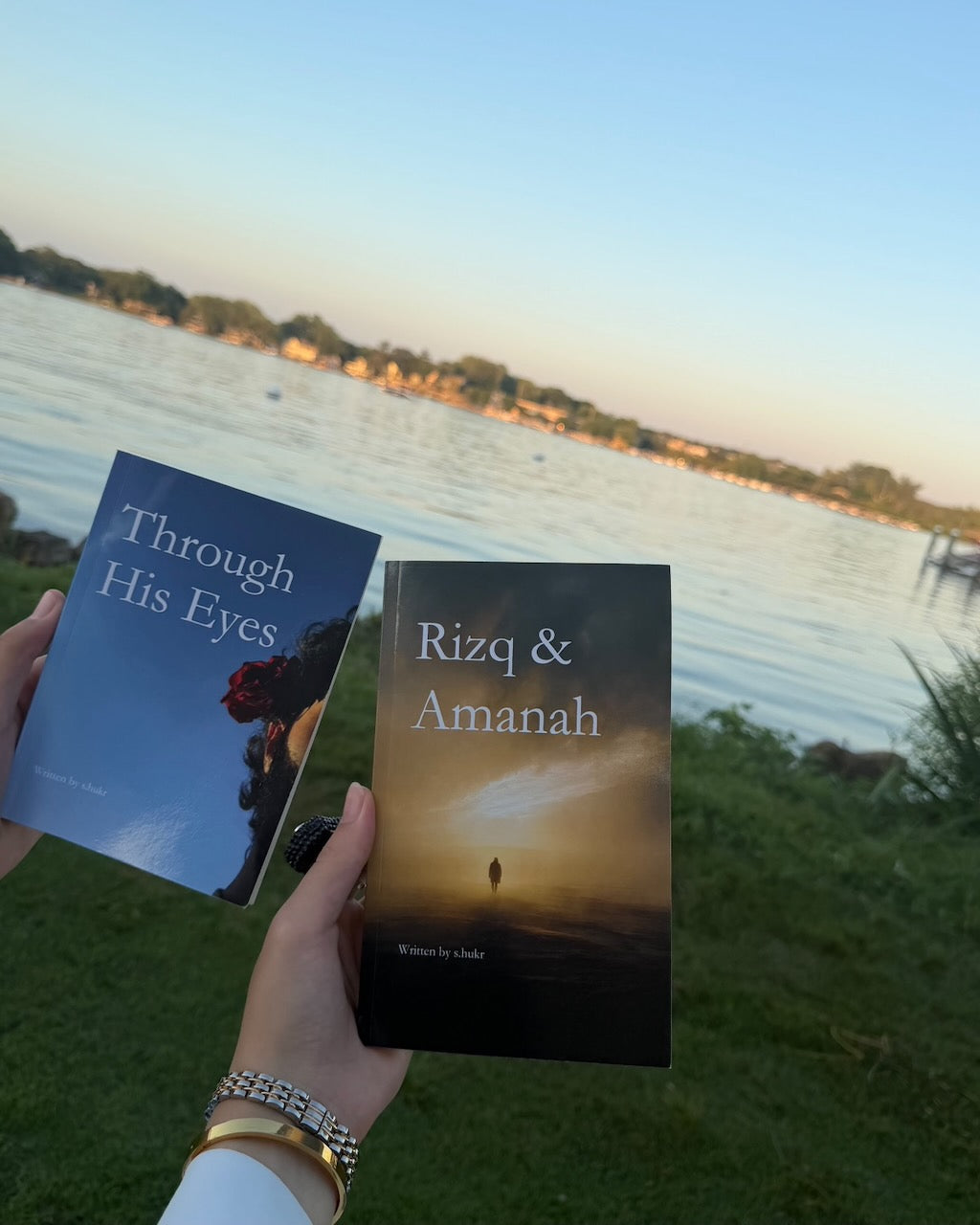 Two books are held up by a person outdoors near a lake at sunset: 'Through His Eyes' on the left with a blue-toned cover and 'Rizq & Amanah' on the right with a dark warm-toned cover.