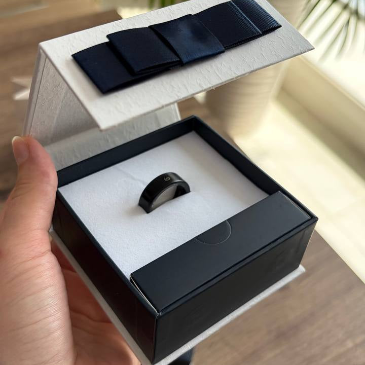 A hand holds an open black and white jewelry box containing a black ring, with a matching black ribbon on the lid, set against a blurred indoor background with a plant.