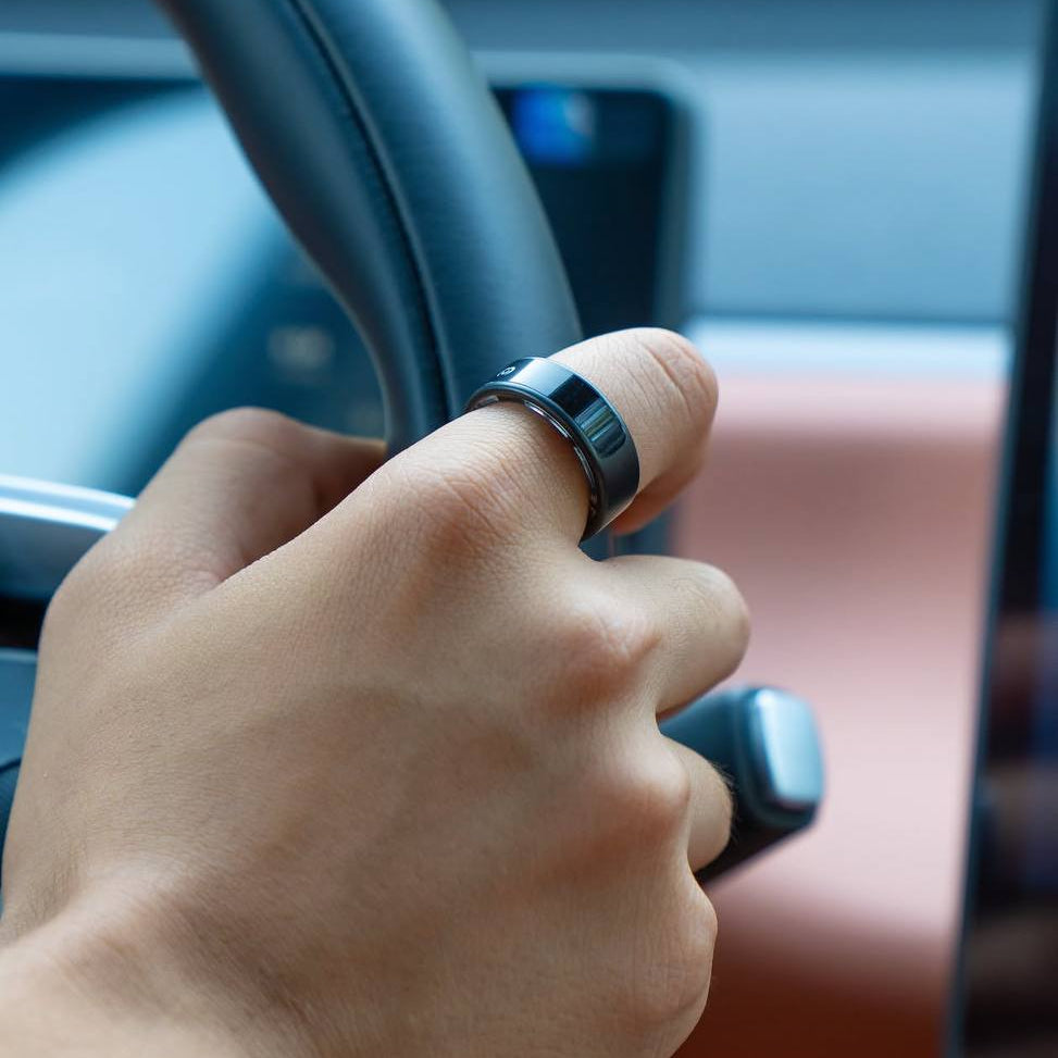 A close-up of a person's hand holding a car steering wheel while wearing a slim black ring with a digital display on the side, inside a vehicle with a brown dashboard.
