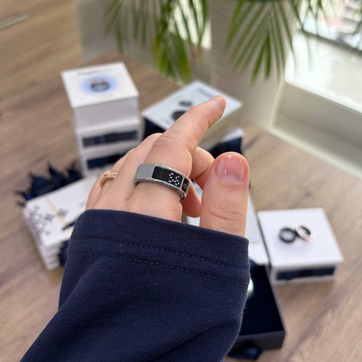 A close-up of a person's hand wearing a silver-toned ring with a black digital display, pointing toward the ring. Several white product boxes are scattered on a wooden table in a bright indoor setting with a green potted plant in the background.