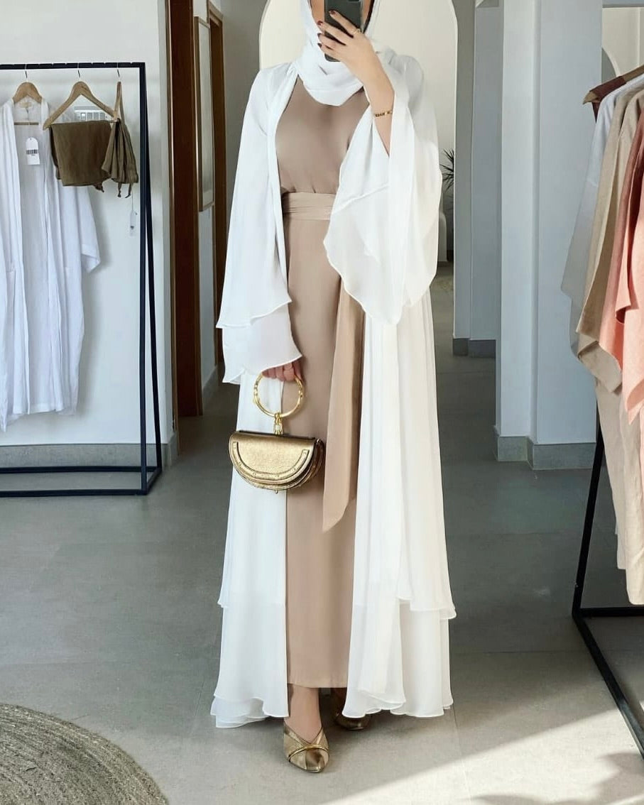 A woman stands in a boutique wearing a beige two-piece dress with a long white draped overlay and a white headscarf. She is holding a gold crescent-shaped handbag and wearing gold flat shoes.